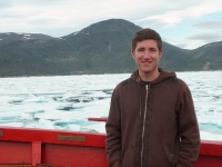 Ian Church stands on the deck of a red vessel with sea ice and hills visible behind him.
